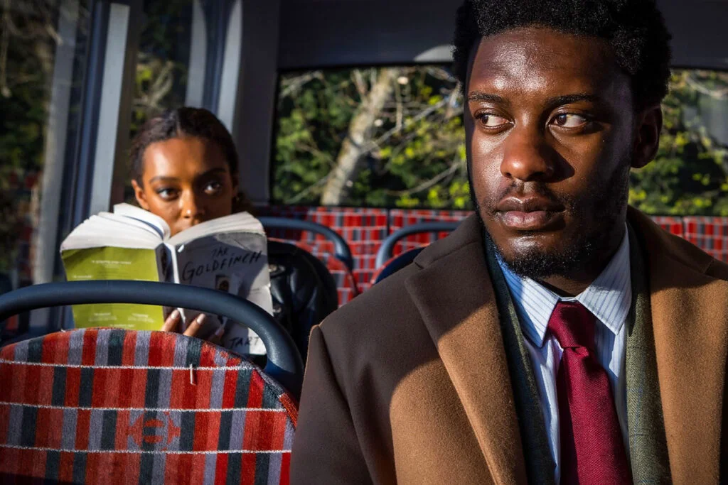Ein Mann mit braunem Mantel und roter Krawatte sitzt im Bus. Hinter ihm eine Frau, die ein Buch liest.