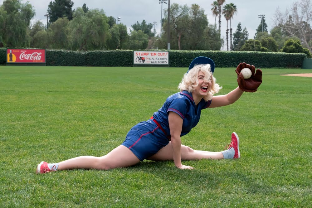 Eine blonde Frau in blauem Sportoutfit macht den Spagat. In der linken Hand hält sie einen Baseballhandschuh und eine Baseball.