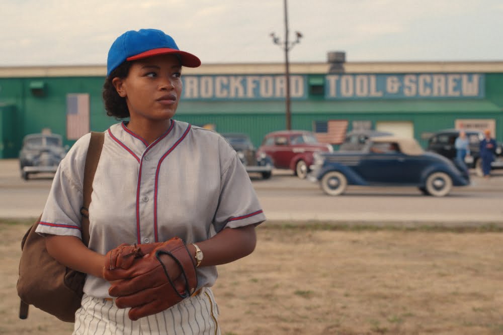 Eine junge Schwarze Frau in Sportkleidung und mit einem Baseballhandschuh. Im Hintergrund alte Autos und ein Fabrikgebäude.