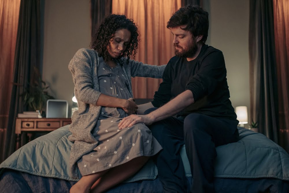 Ein Paar sitzt auf einem Bett. Die schwangere Frau hält eine Hand auf ihren Bauch.