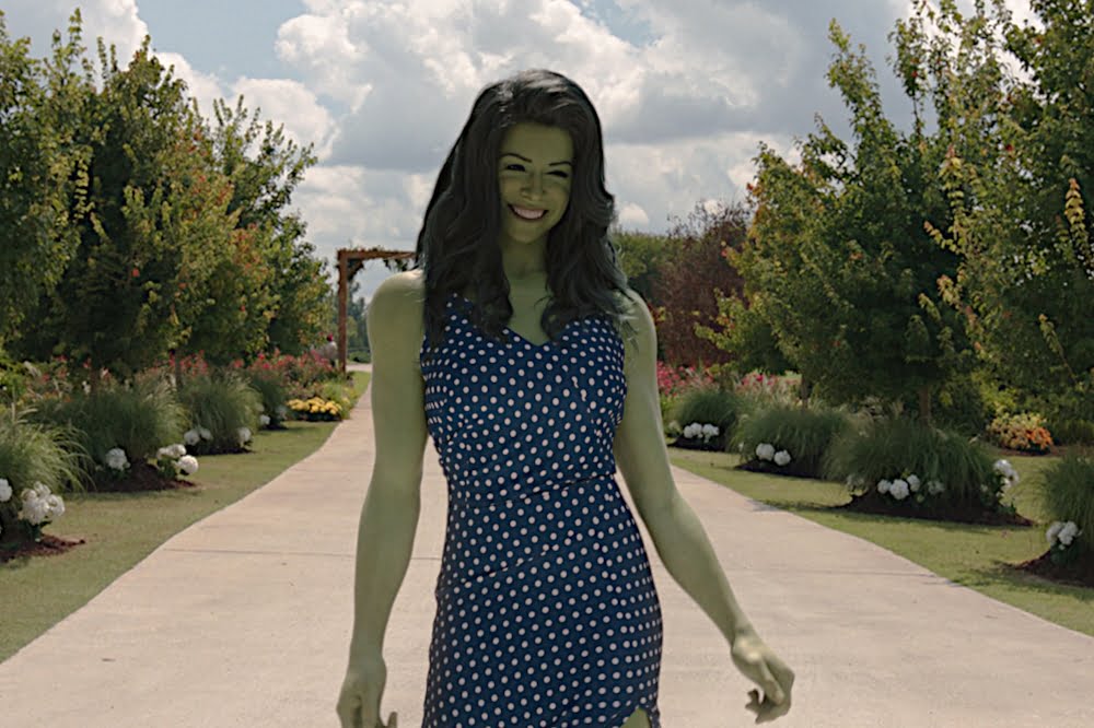 Eine Frau im blauen Kleid mit weissen Punkten, lange schwarze Haare und grünliche Haut steht auf einem Weg umrahmt von kleinen Bäumen.
