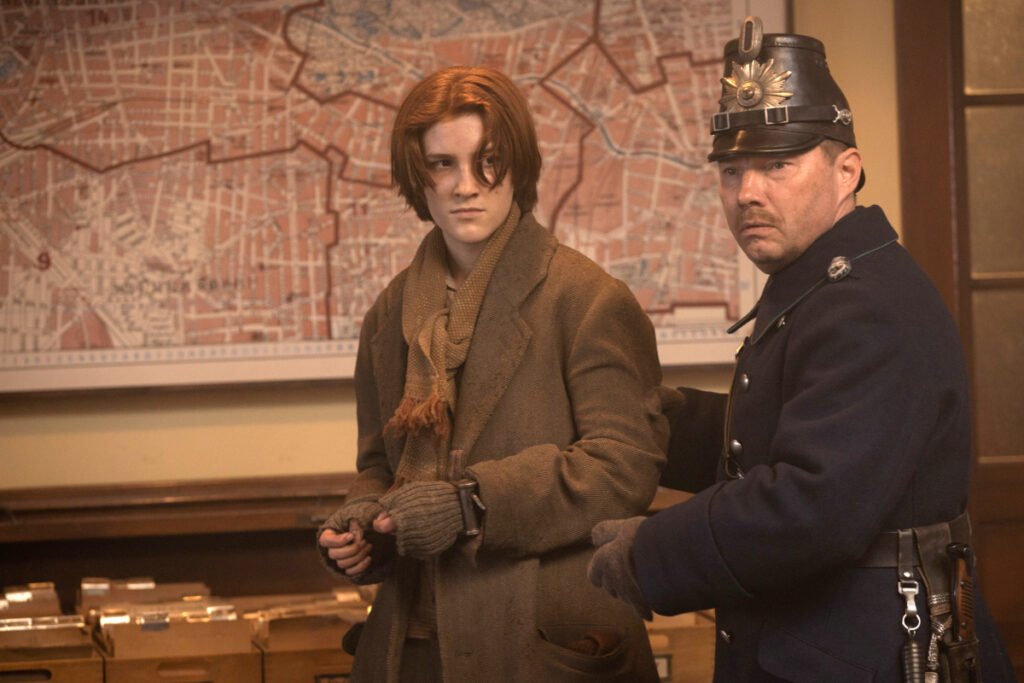 Eine junge rothaarige Frau in einem schäbigen Mantel und in Handschellen wird von einem Polizisten abgeführt. Im Hintergrund eine Landkarte. 