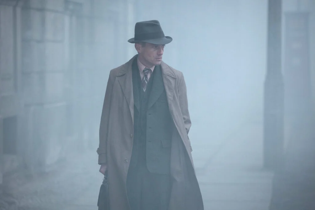 Ein Mann mit Hut, Anzug und hellbraunem Mantel im Nebel.