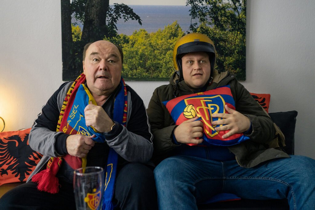 Zwei Männer sitzen auf einer Couch. Sie tragen Fan-Utensilien des FC Basel.