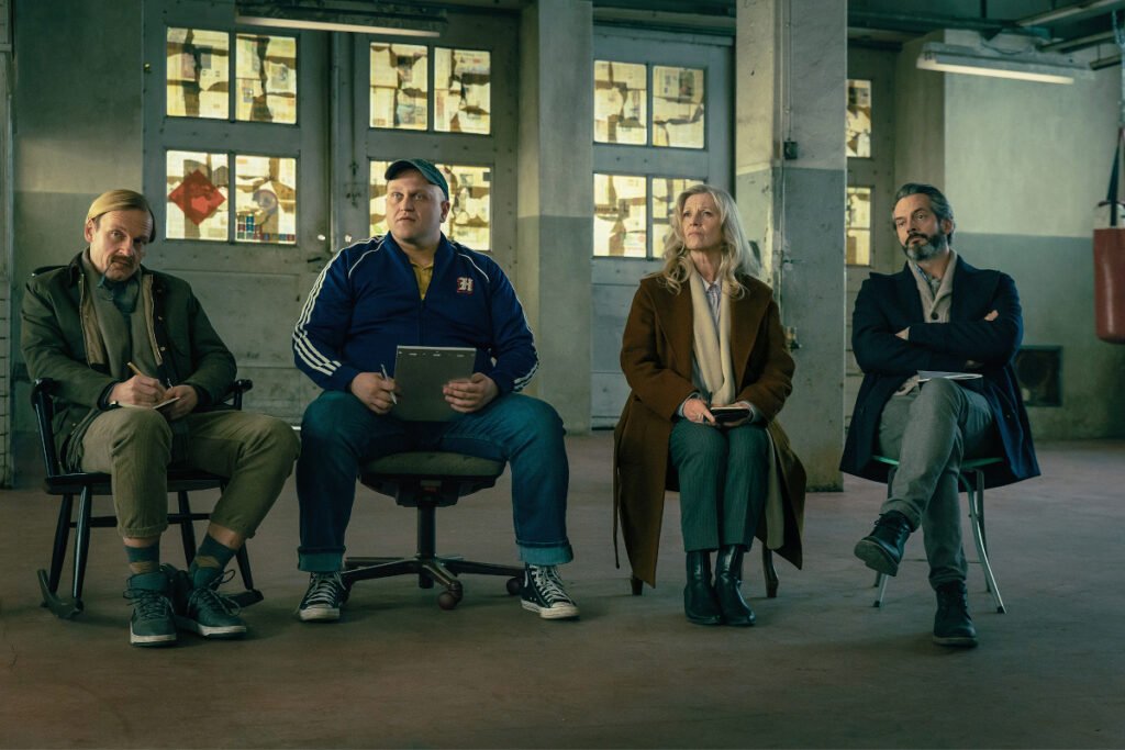 Drei Männer und eine Frau sitzen in einer Art Fabrikhalle auf Stühlen.