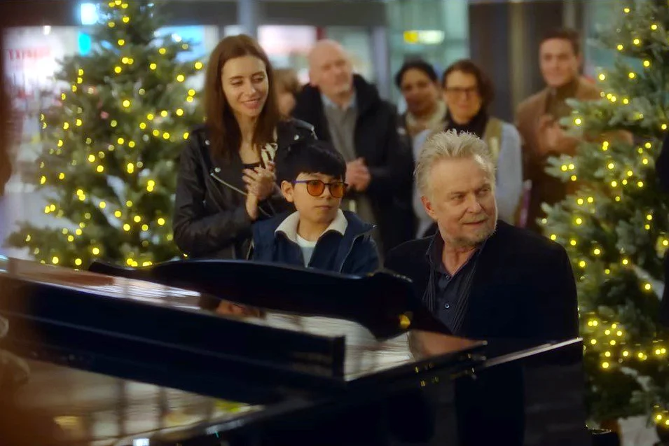 Ein Mann sitzt am Klavier. Hinter ihm stehen Zuschauer:innen. Zwei Weihnachtsbäume umrahmen die Szene.