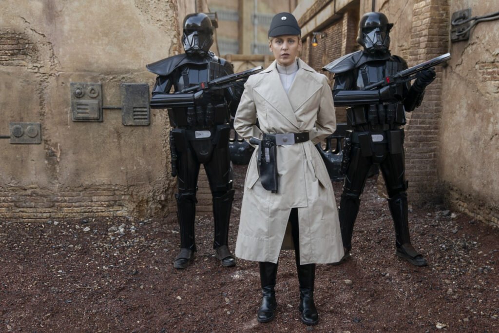 Eine Frau in Uniform, weisser Mantel und schwarze Stiefel. Hinter ihr zwei Stormtrooper in schwarz. 