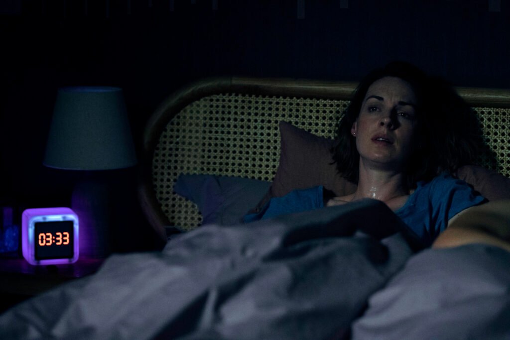Eine Frau liegt wach im Bett. Der Wecker auf dem Nachttisch zeigt 3.33 Uhr an. 
