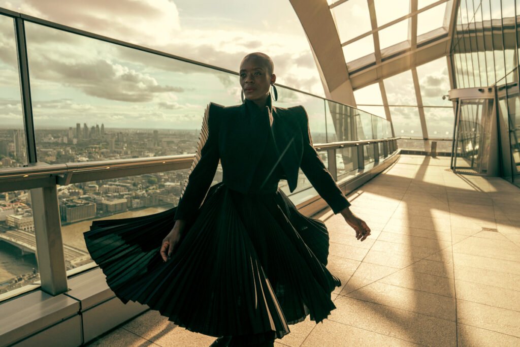 Eine Frau steht auf einer Terrasse hoch über einer Stadt. Sie trägt ein futuristisches, wehendes schwarzes Kleid.