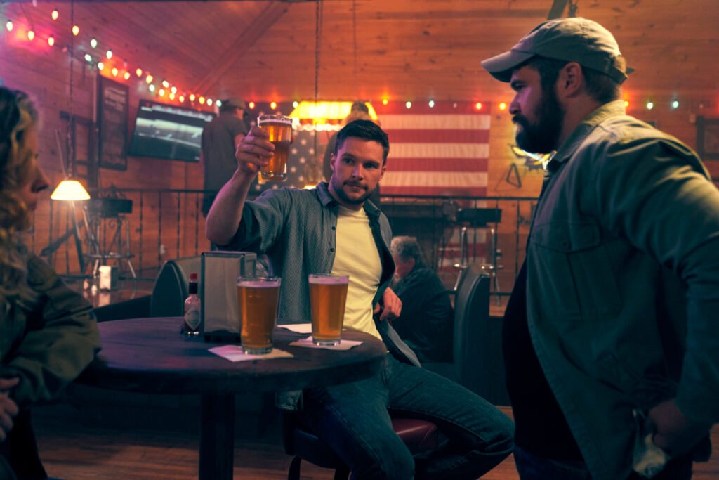 Zwei Männer in einer Bar. Auf dem Tisch stehen zwei Biergläser. Einer der Männer hat sein Glas erhoben.