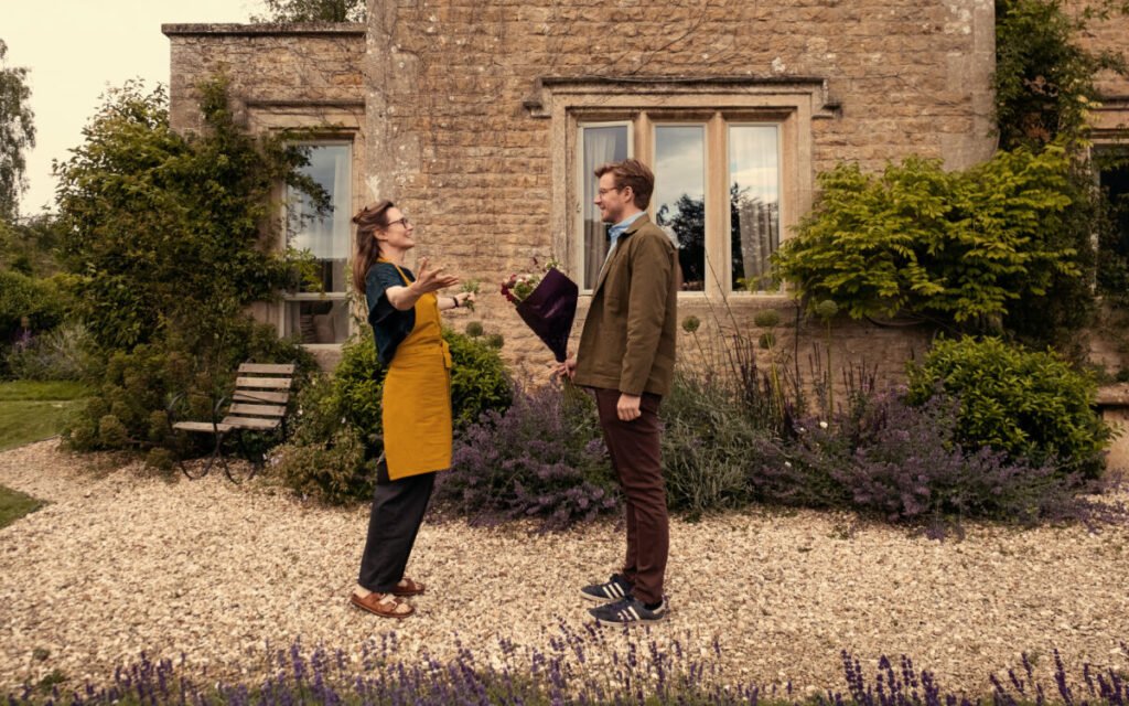 Ein Mann und eine Frau auf einem Kieselweg vor einem Haus. Er hält einen Blumenstrauss in der Hand. Sie begrüsst ihn mit offenen Armen. 