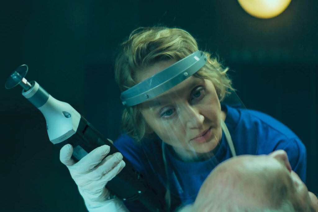 Eine Frau mit Gesichtsschutz und einem Werkzeug in der Hand blickt auf den Kopf einer Leiche.