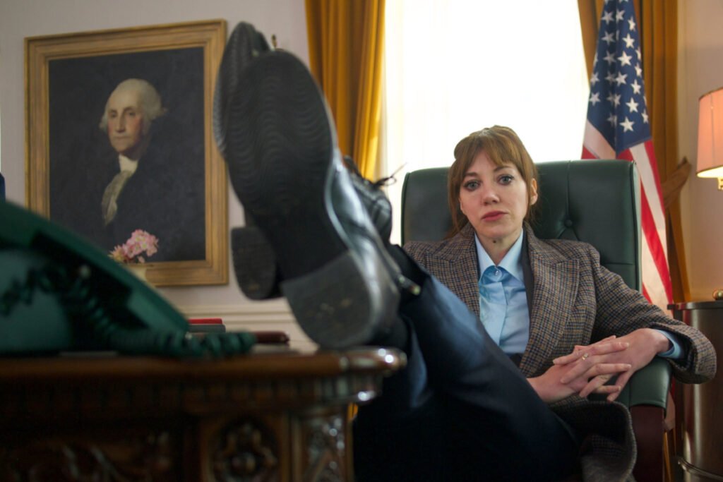 Eine Frau sitzt in einem Ledersessel im Oval Office und streckt die Beine auf den Schreibtisch. Im Hintergrund die US-amerikanische Flagge und ein Ölgemälde von George Washington.