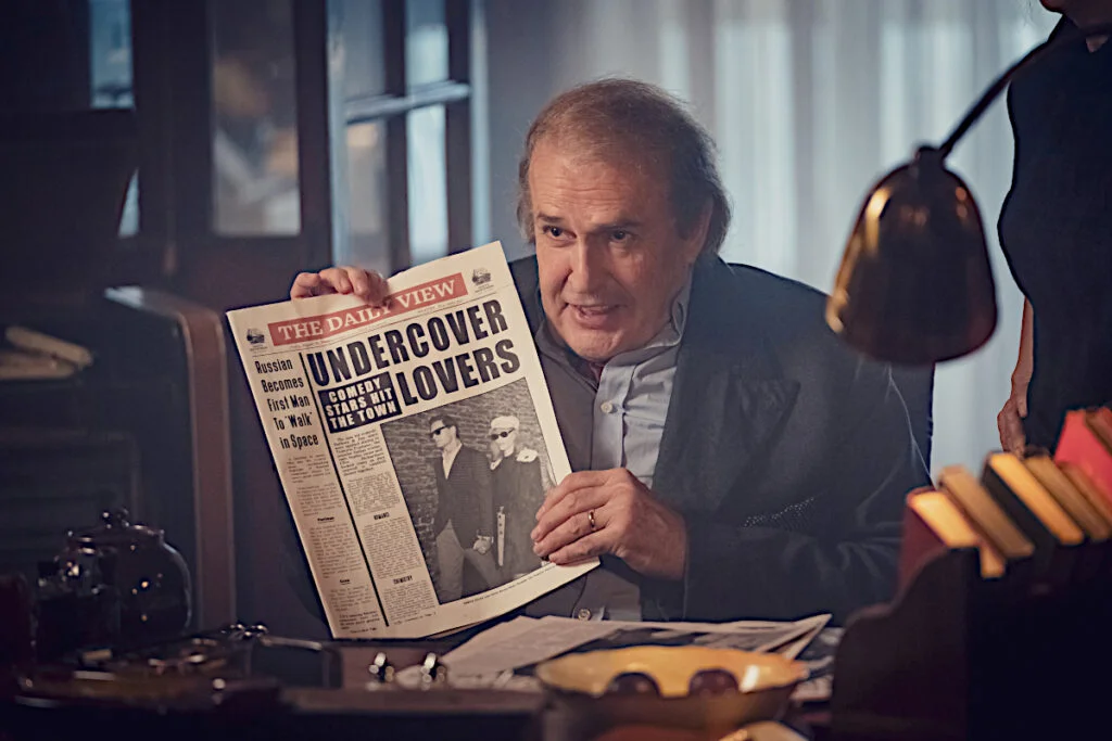 Ein Mann sitzt an einem Schreibtisch und zeigt das Titelblatt einer Zeitung. 