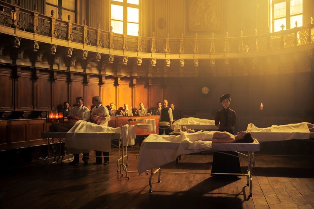 Ein halbrunder Raum. Vier Leichen liegen auf Seziertischen. Eine Frau steht hinter einem dieser Tische. Männer beobachten die Arbeit von zwei Ärzten an einem anderen Tisch.