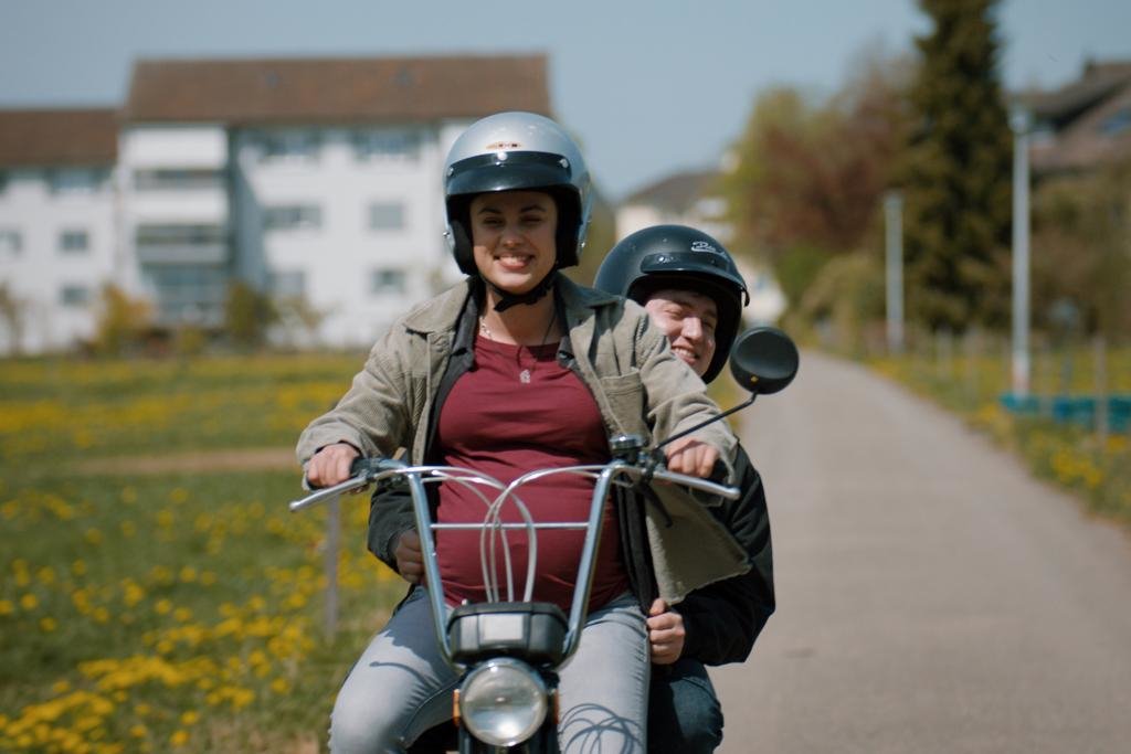 Eine schwangere Frau mit Helm auf einem Moped. Auf dem Gepäckträger sitzt auch noch ein Mann. 
