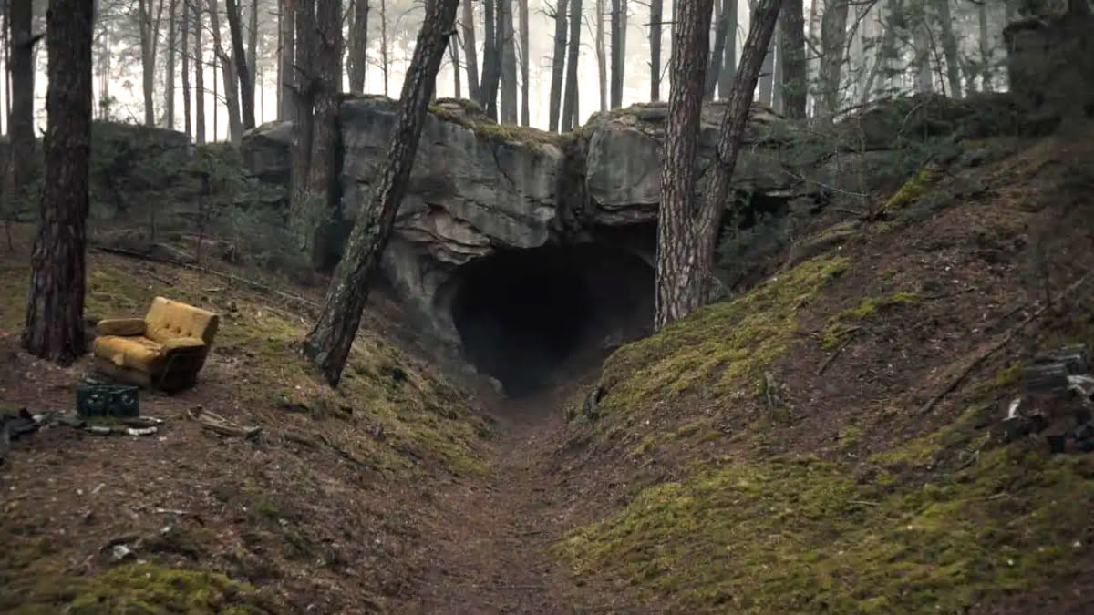 In einem Wald führt ein Trampelpfad zum Eingang zu einer Höhle. Am Rand steht ein Sofasessel. 