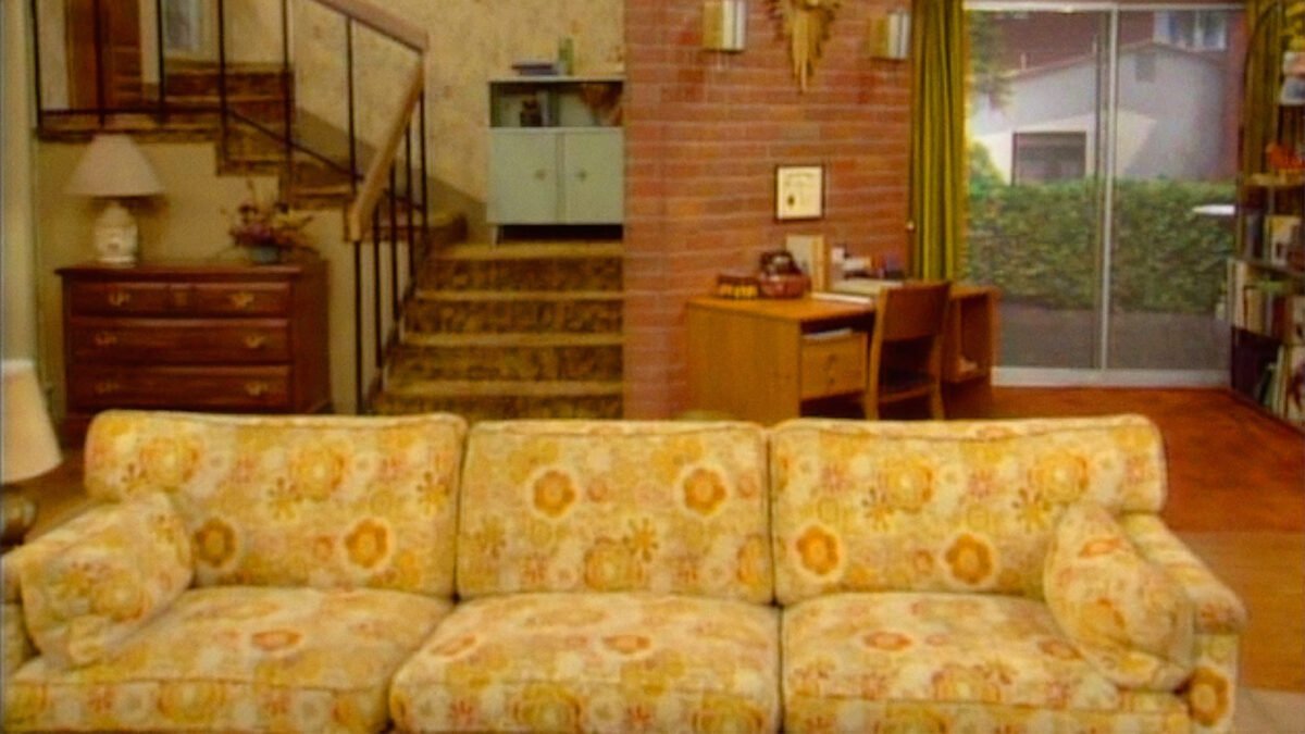 Eine gelbe Couch steht in einem Wohnzimmer. Eine Treppe führt in den ersten Stock.
