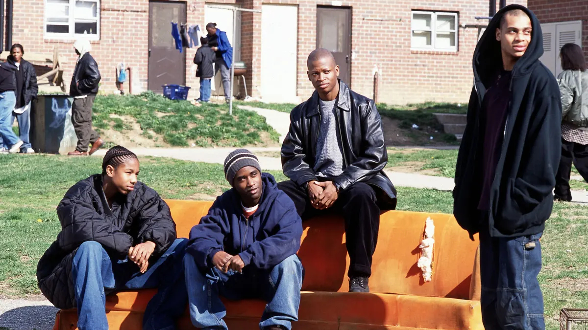 Vier Schwarze Jugendliche in einem Hof. Drei sitzen auf einer orangen Couch, die auf dem Gras steht.