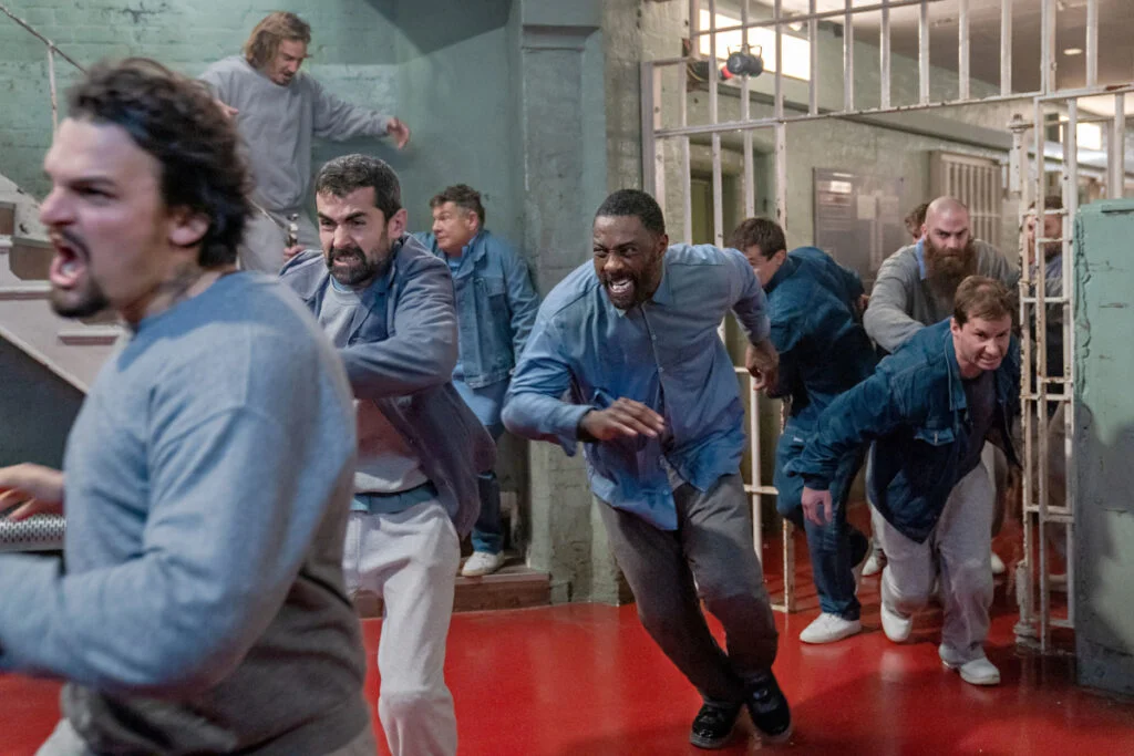 Männer in einem Gefängnis, die mit verzerrten Gesichtern durch eine geöffnete Gittertür rennen.