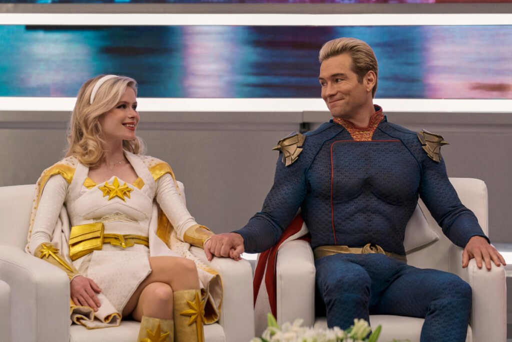 Ein Mann und eine Frau in Superheldenkostümen sitzen in einem Fernsehstudio. Sie halten sich die Hände und schauen sich lächelnd an.