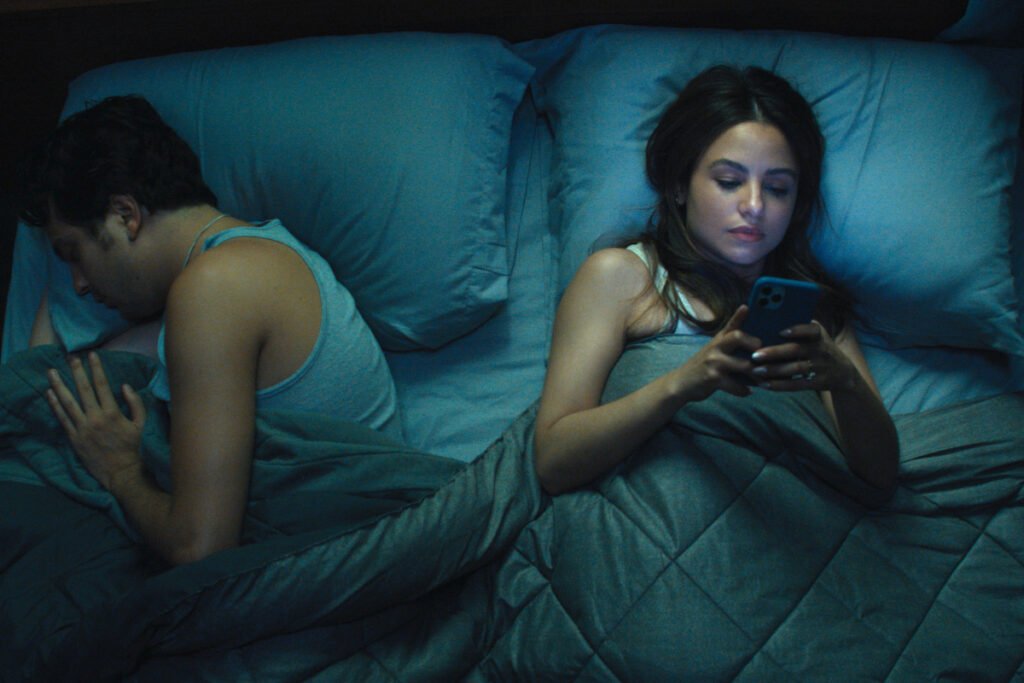 Ein Mann und eine Frau liegen im Bett. Er schläft. Sie hält ein Smartphone in der Hand und schaut auf den Bildschirm.