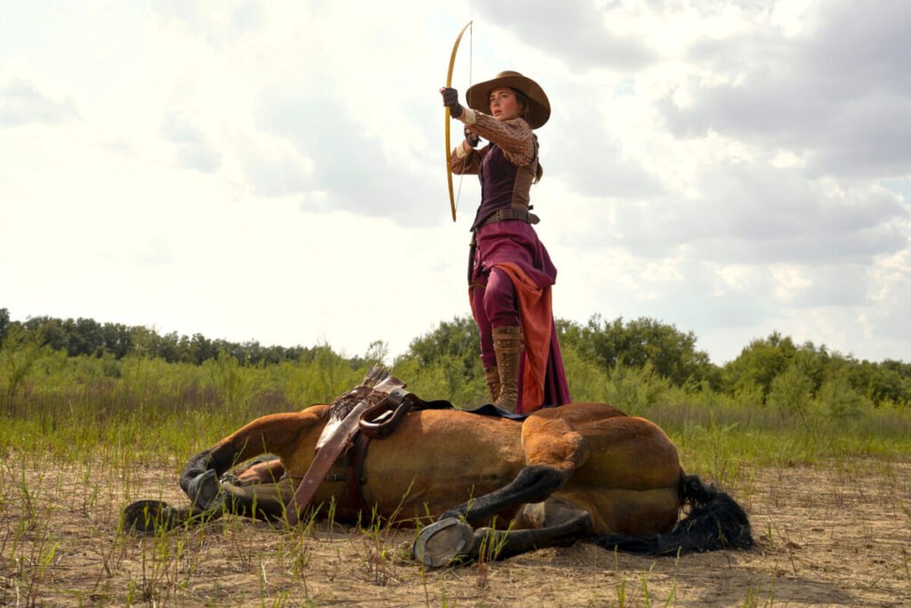 Eine Frau hat einen Fuss auf einem toten Pferd abgestützt und hält einen gespannten Pfeilbogen in der Hand.