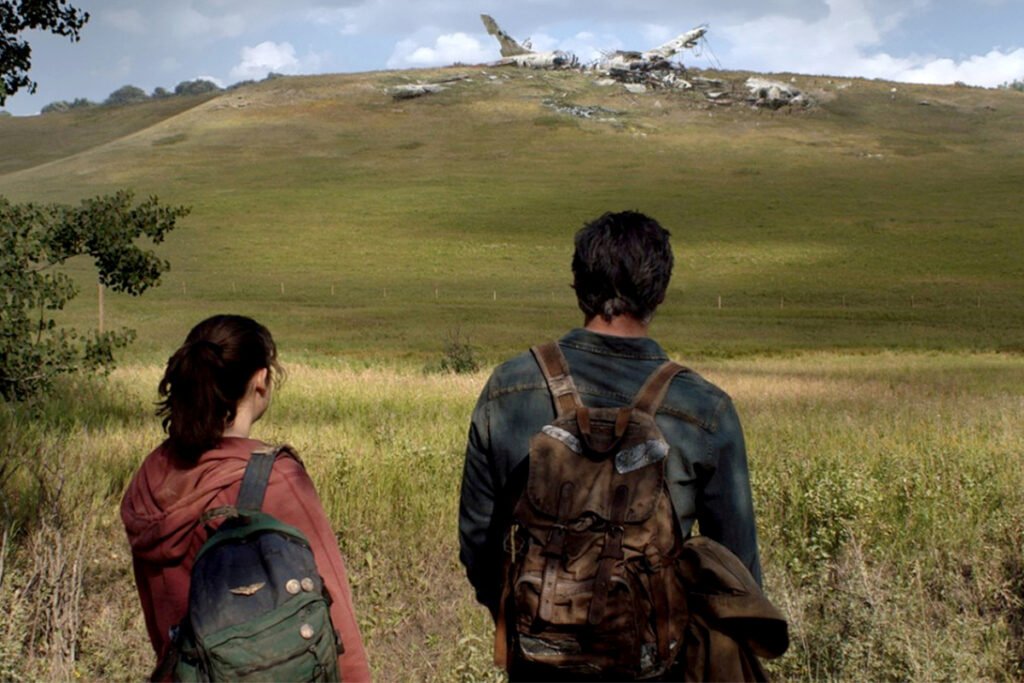 Eine junge Frau und ein Mann sehen in der Ferne auf einem Hügel ein abgestürztes Flugzeug.
