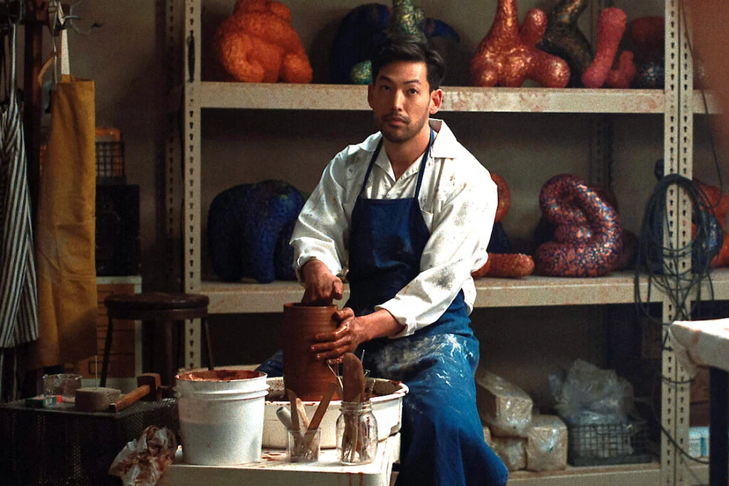 Ein Mann sitzt in einer Werkstatt an einer Töpferscheibe. Im Hintergrund ein Regal mit vielen ähnlichen Skulpturen.
