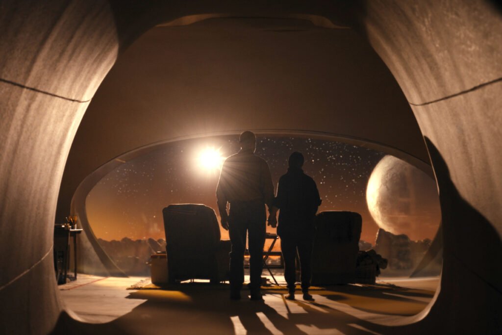 Eine Frau und ein Mann stehen in einem kuppelartigen Raum in dem zwei Sessel stehen. Ein ovales Fenster gibt den Blick frei auf einen fremden Planeten.