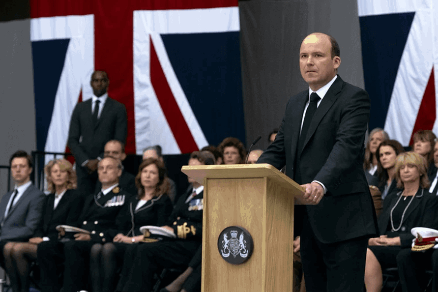 Ein Mann an einem Rednerpult. Britische Flaggen im Hintergrund. 