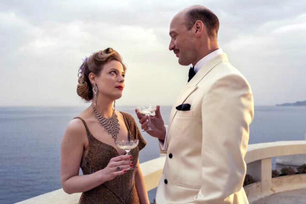 Eine Frau und Mann in eleganten Kleidern stehen mit Gläsern in der Hand auf einer Terrasse, im Hintergrund das Meer.