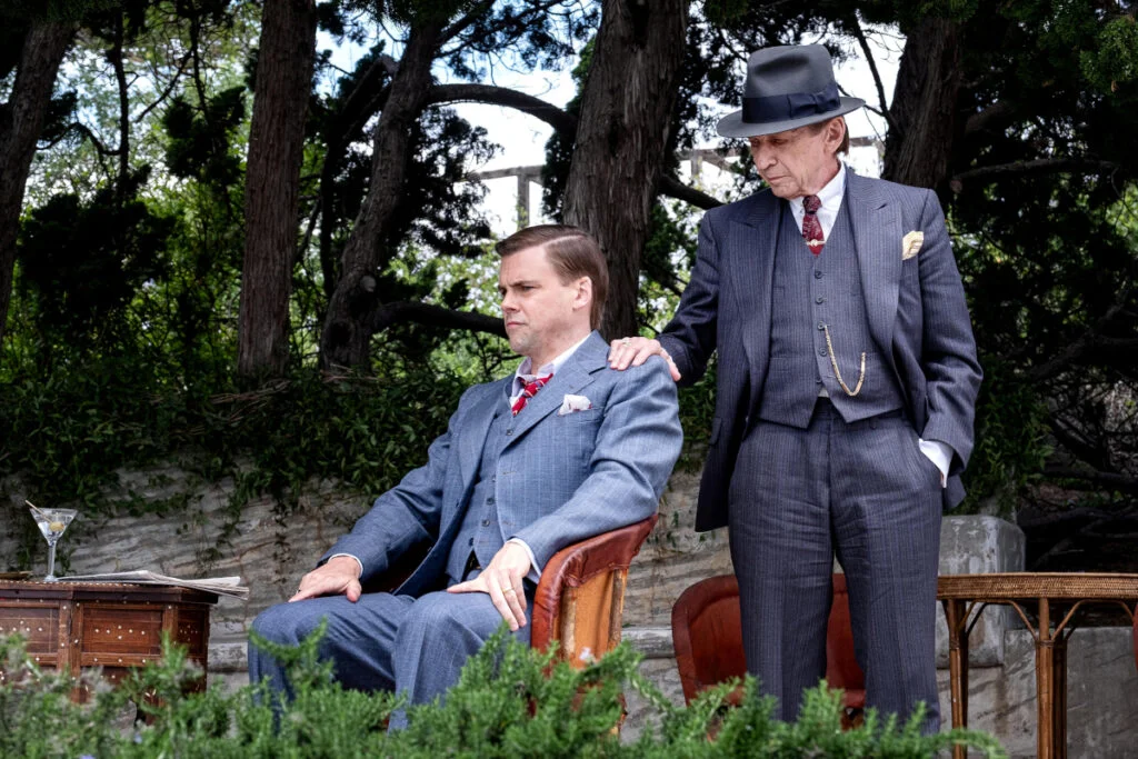 Ein Mann sitzt in einem Sessel in einem Garten. Ein älterer Mann steht hinter ihm, die Hand auf seine Schulter gelegt.