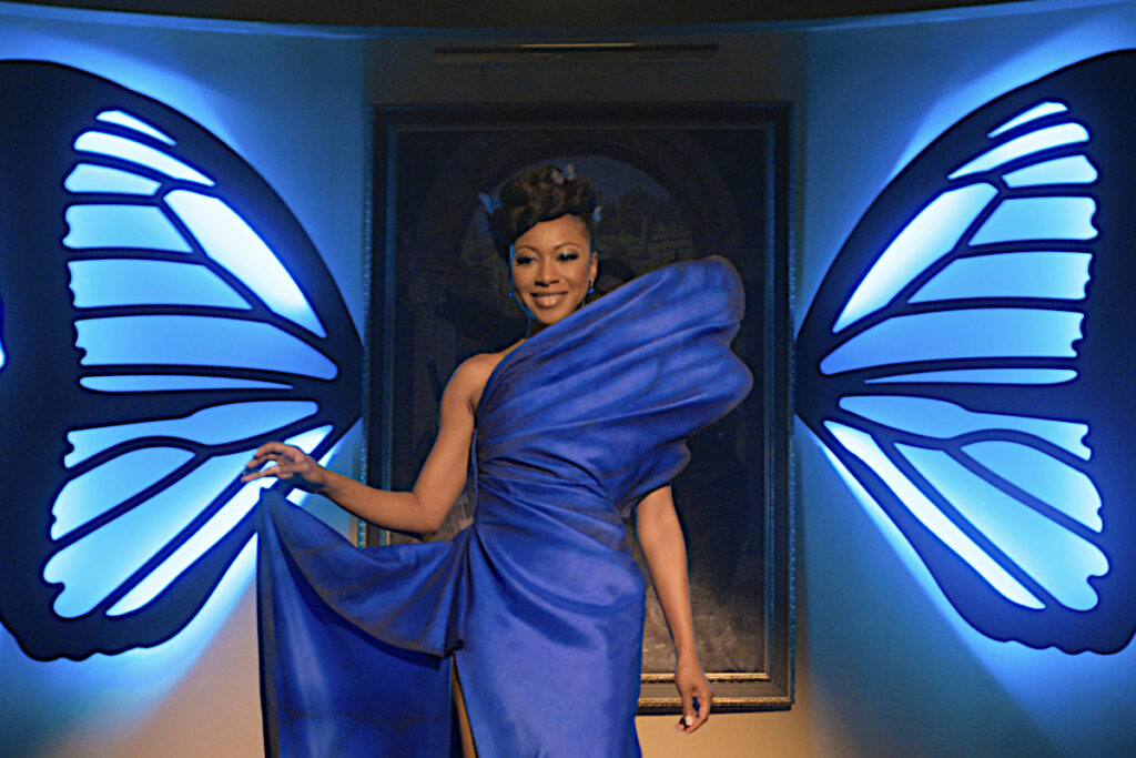 Eine Frau in einem extravaganten blauen Abendkleid, die durch eine Tür schreitet, die mit Schmetterlingsflügel verziert ist.