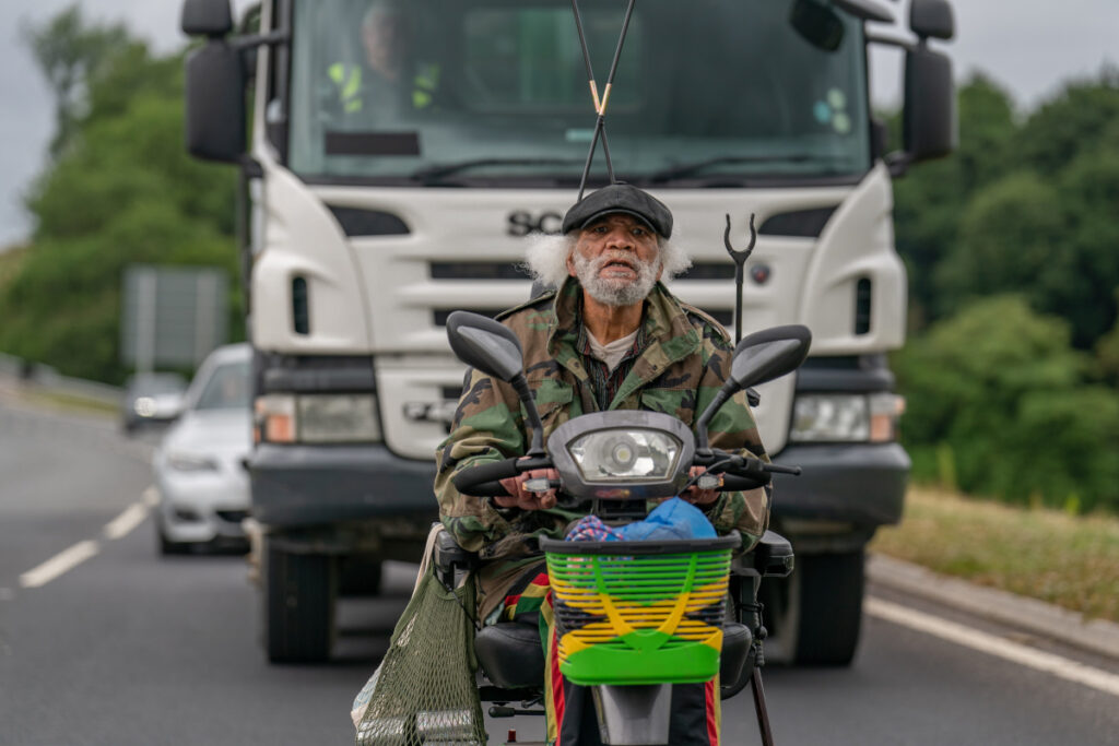 Ein älterer Mann auf einem Elektroscooter. Dahinter ein Lastwagen und weitere Autos.