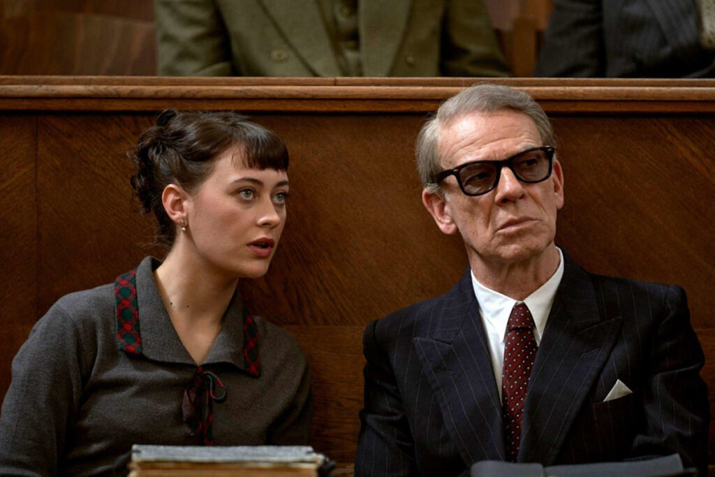 Eine junge Frau und ein älterer Mann mit dunkler Brille sitzen als Zuschauer auf einer Bank im Gerichtssaal.