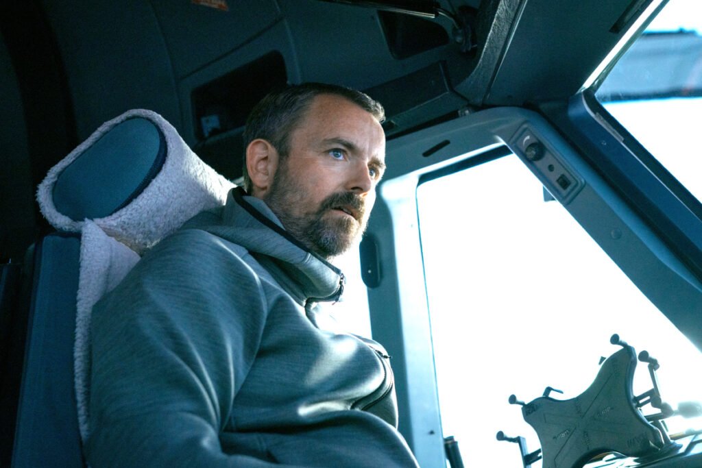 Ein Mann mit grauem Kapuzenpullover sitzt im Cockpit im Pilotensessel.