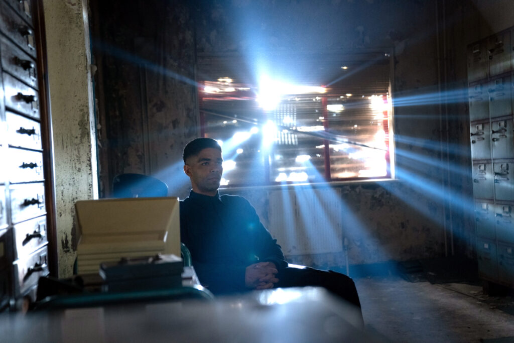 Ein Mann sitzt in einem heruntergekommenen Raum. Durch das mit Brettern zugenagelte Fenster kommen vereinzelt Lichtstrahlen durch.