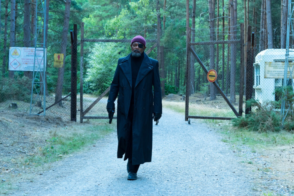 Ein Mann im langen Mantel mit Mütze und Augenklappe, Pistole in der Hand, geht auf einem Weg in einem Wald. Im Hintergrund ein hoher Zaun.