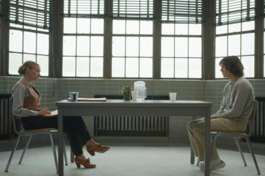 Eine Frau und ein Mann sitzen an den Kopfenden eines Tisches vor einer Fensterfront.