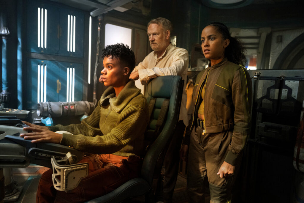 Zwei junge Frauen und ein älterer Mann stehen im Cockpit eines Raumschiffs und schauen staunend nach vorn.