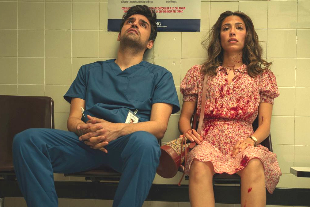 Eine Frau und ein Mann sitzen auf einer Bank. Er trägt eine Krankenpflegeruniform, sie ein Kleid mit Blutflecken.