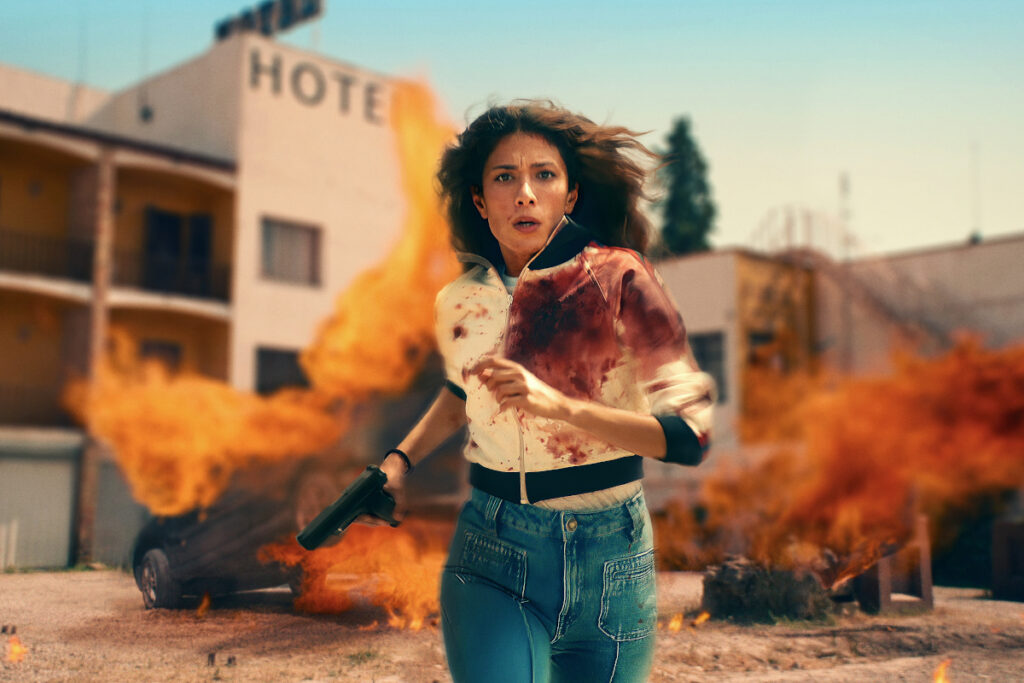 Eine Frau in blutverschmierten Kleidern rennt von einem Gebäude weg, vor dem Autos in Flammen stehen.