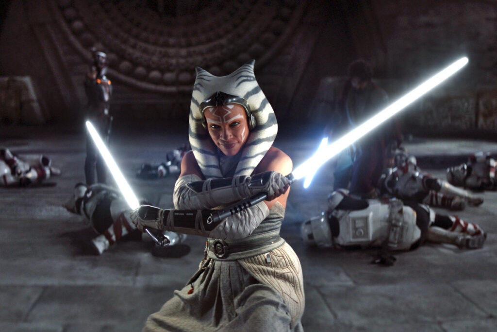 Die Jedi-Ritterin Ahsoka mit gezücktem Lichtschwert in jeder Hand. Im Hintergrund am Boden Stormtroopers.