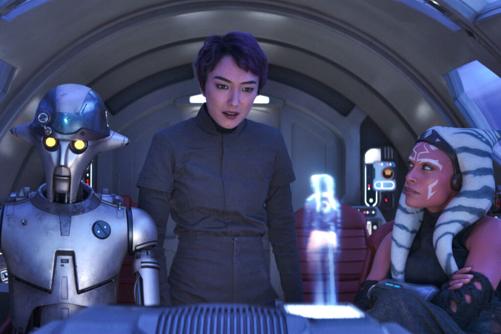 Ein Droide und zwei Frauen im Cockpit eines Raumschiffs. Sie schauen auf die Projektion einer weiteren Figur, die klein im Vordergrund zu sehen ist.