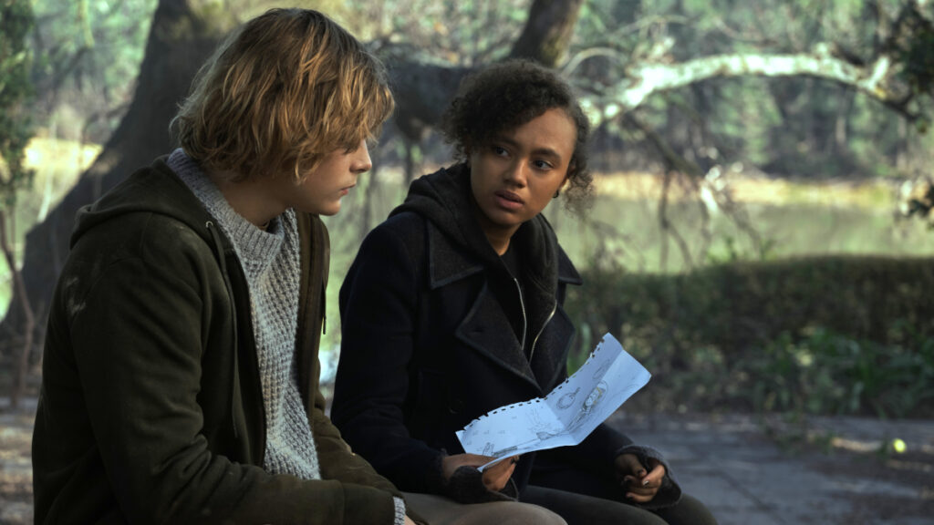 Zwei Jugendliche sitzen auf einer Bank. Die junge Frau zeigt dem jungen Mann ein Blatt Papier.