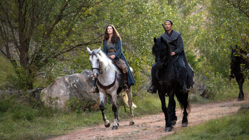 Eine Frau auf einem weissen Pferd, ein Mann auf einem schwarzen Pferd reiten durch einen Waldweg.