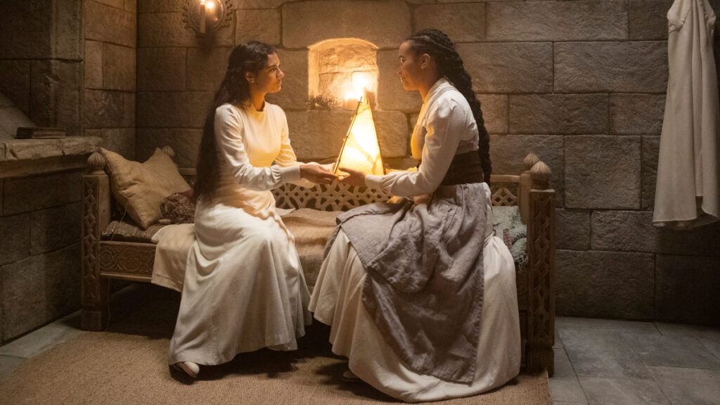 Zwei Frauen in weissen Gewändern sitzen in einer Kammer mit Steinwänden. Sie halten eine zylinderförmige Lampe in den Händen.
