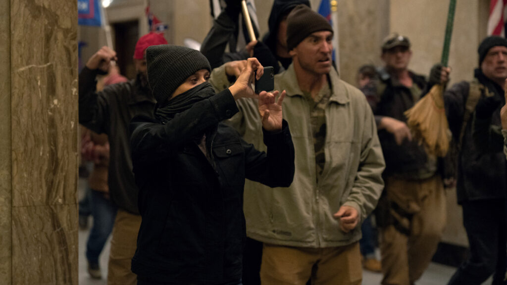 Eine Frau mit Wollmütze und Maske filmt mit ihrem Handy umgeben von mehreren Protestierenden. 