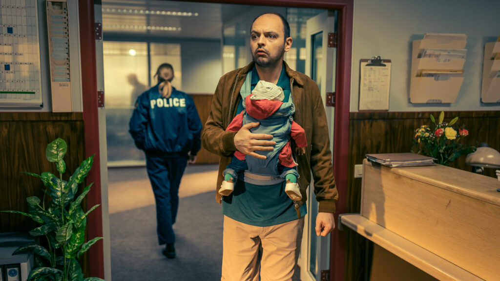 Ein Mann in einer Polizeistation. Er hat ein Baby in einem Tragegestell umgehängt.